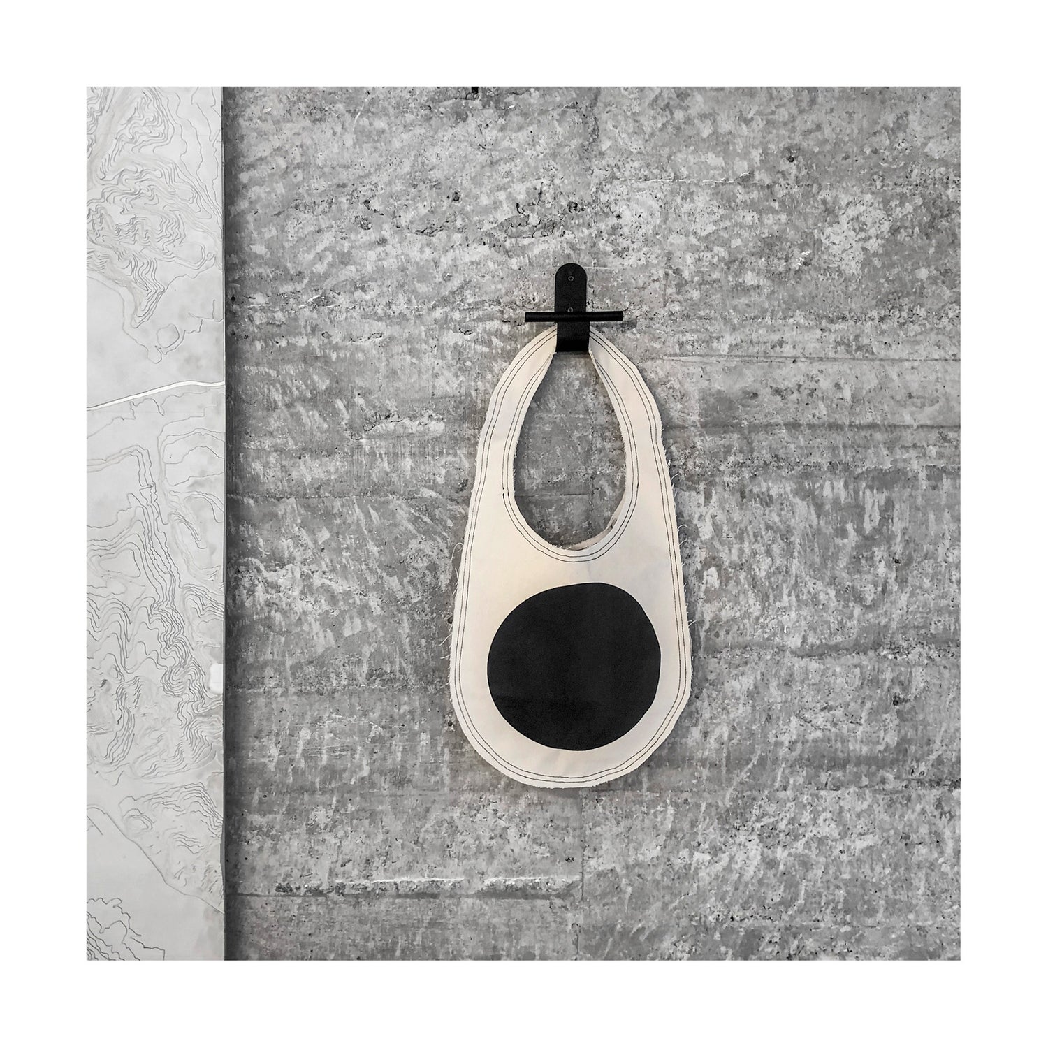 imagem quadrada mostra bolsa tiracolo grande de formato oval, com círculo preto grande estampado no centro, pendurada em cabideiro de aço preto fixado numa parede de concreto